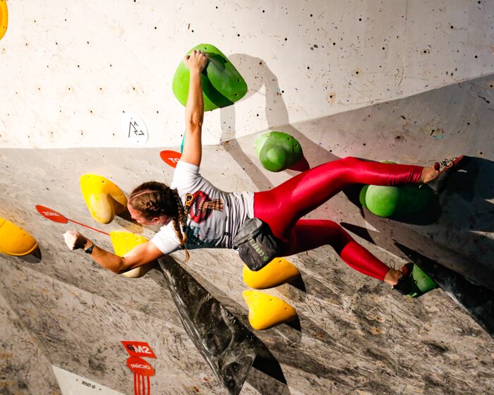 atleta em movimento numa via de boulder da Toca, o corpo estendido entre uma agarra verde no alto e os apoios amarelos