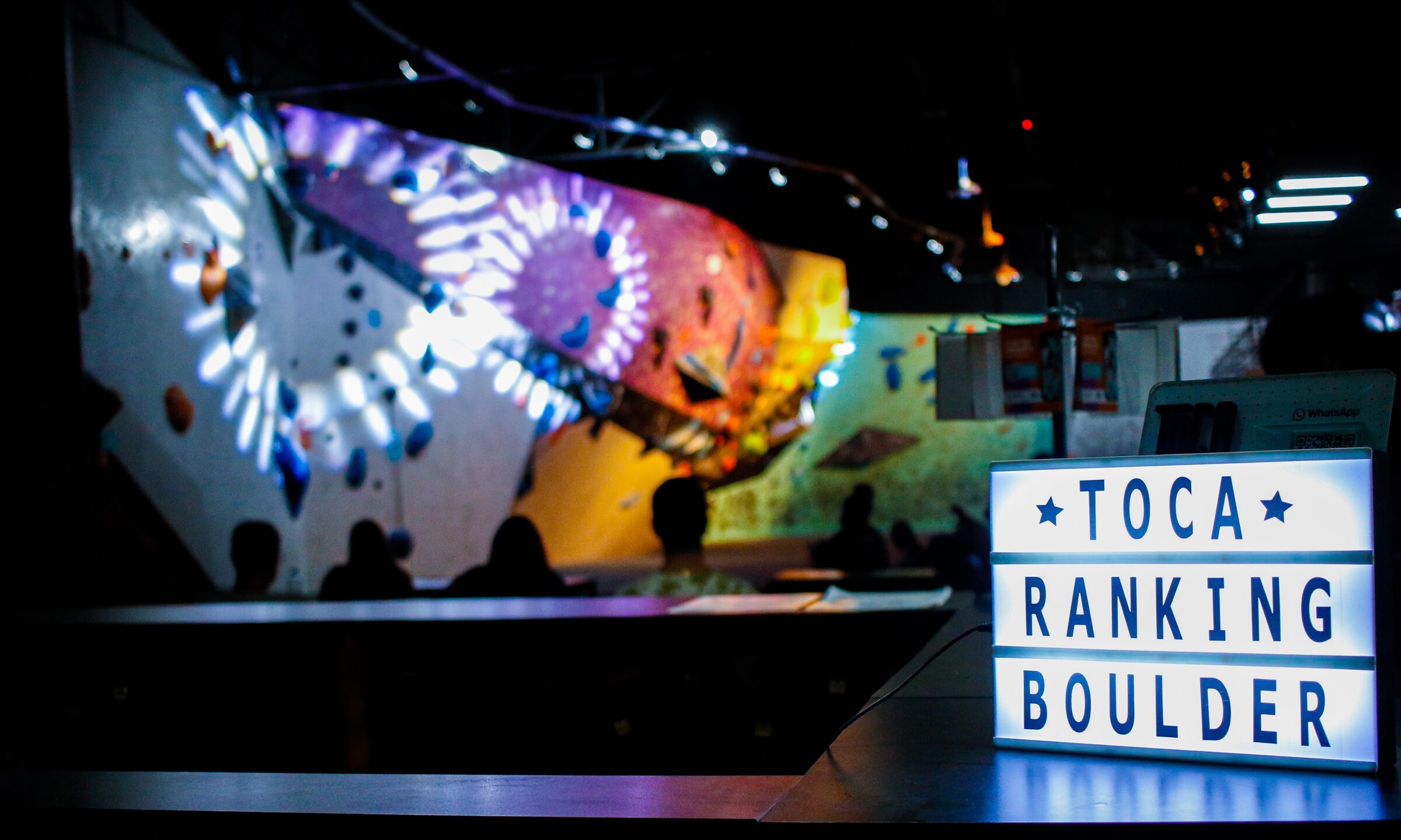 o light box do TOCA RANKING BOULDER aceso no balcão da Toca, com a galera assistindo à parede de boulder iluminada ao fundo
